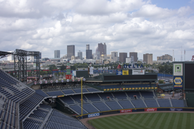 Eine wolkenverhangene Sicht auf ein Baseballstadion mit leeren Sitzplätzen, umgeben von einer Stadtkulisse, Bäumen und fernen Gebäuden.