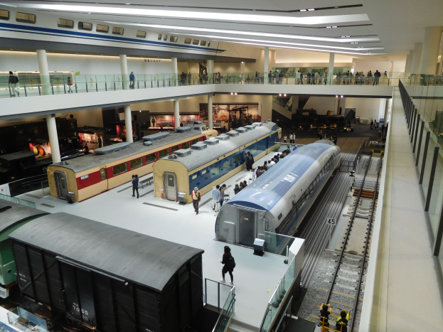 Ein großer Museumsraum mit verschiedenen Zügen in unterschiedlichen Größen und Farben, mit Menschen, Geländern, Säulen und Deckenleuchten.