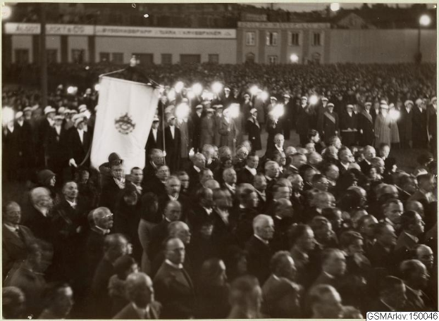 Schwarzes und weißes Bild einer großen Menge vor einem Gebäude, mit einer Person, die eine Fahne in der Mitte hält, Stangen und Lichter im Hintergrund und Text unten, der "Gustav Klimts Rede an der Berliner Mauer" lautet.
