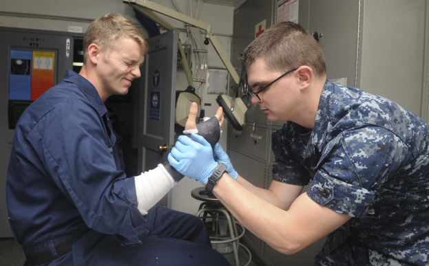 Zwei Männer in Militäruniformen, einer sitzt in einem Stuhl mit einem Gips am Arm und der andere steht neben ihm, in einer medizinischen Einrichtung mit verschiedenen Geräten im Hintergrund.