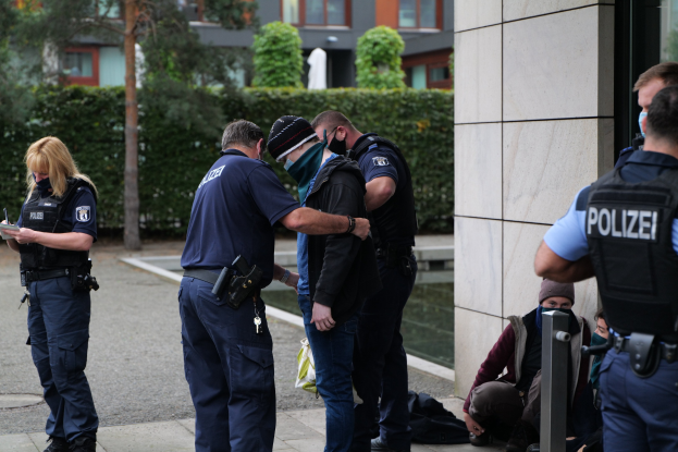 Gruppe von Polizeibeamten vor einem Gebäude mit einer Frau, die ein Buch und einen Stift hält, sowie anderen Personen in der Nähe, umgeben von Grünflächen und städtischen Strukturen.