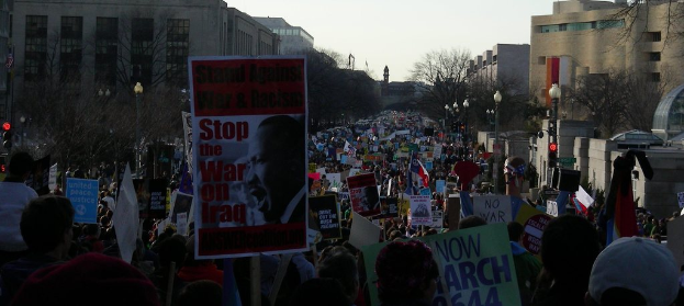 Eine Gruppe von Menschen demonstriert auf einer Straße und hält Plakate, mit Gebäuden, Bäumen, Laternenpfählen und dem Himmel im Hintergrund.