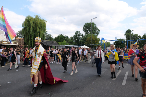 Eine Gruppe von Menschen, die bei der Gay Pride Parade 2018 marschieren und eine Regenbogenflagge sowie Musikinstrumente tragen, mit Laternenpfählen, Bäumen, Hütten und einem bewölkten Himmel im Hintergrund.