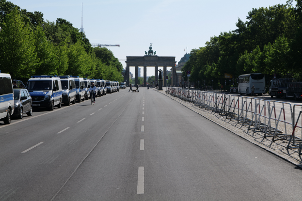 Eine Reihe von Polizeiwagen, die auf der Seite einer Straße vor dem Brandenburg-Tor in Berlin, Deutschland, geparkt sind, mit Menschen, die Fahrräder fahren und auf der Straße stehen, Barrieren und Bäume, die die Seiten säumen, und ein Tor mit Statuen im Hintergrund.