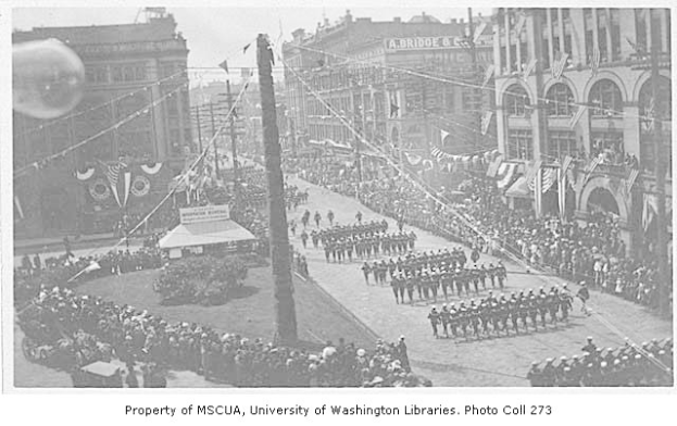 Ein Schwarz-Weiß-Foto eines Umzugs auf einer Stadtstraße mit vielen Menschen, einige halten Fahnen, und einem Mast im Vordergrund, mit Gebäuden und Text unten auf dem Bild.
