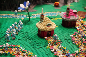 Ein Lego-Modell einer Kleinstadt mit Häusern, Blumen, Spielzeugen, Pflanzen und anderen Gegenständen vor einem leicht unscharfen, träumerischen Hintergrund.