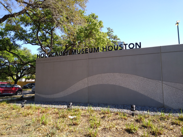 Außenansicht des Houston Holocaust Museums mit eingravierter Textwand, Gedenksteine, Grünfläche, Straßenverkehr, einer Straßenlaterne und einem bewölkten Himmel.