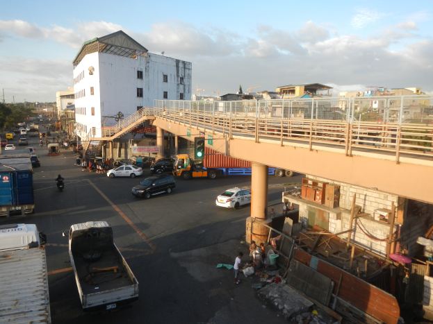 Eine belebte Stra├če mit Autos, Lastwagen, Füßg├Ąngern, einer Brücke mit Gel├Ąndern und Pfeilern, Ampeln an Masten, Geb├Ąude mit Fenstern, B├Ąumen und einem bew├Âlktem Himmel.