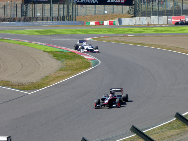Eine Gruppe von Rennwagen fährt auf einer Rennstrecke, umgeben von Gras auf beiden Seiten, einem Maschendrahtzaun mit Bannern und Pfosten im Hintergrund und einigen Zuschauern.