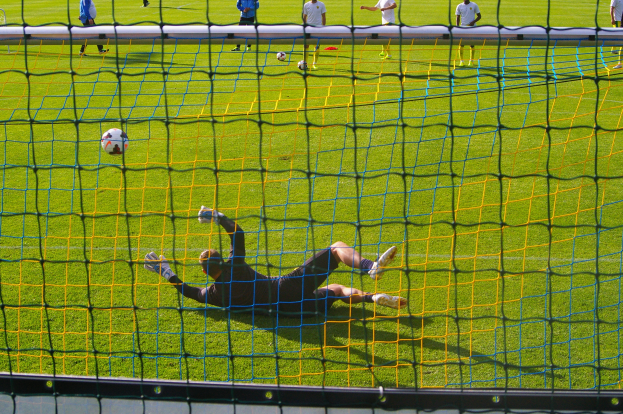 Ein Fußballspieler taucht ab, um einen Ball vor dem Tor zu halten, mit einem Netz im Vordergrund und einigen Menschen im Hintergrund, während mehrere Fußballschuhe auf dem Boden verstreut sind.