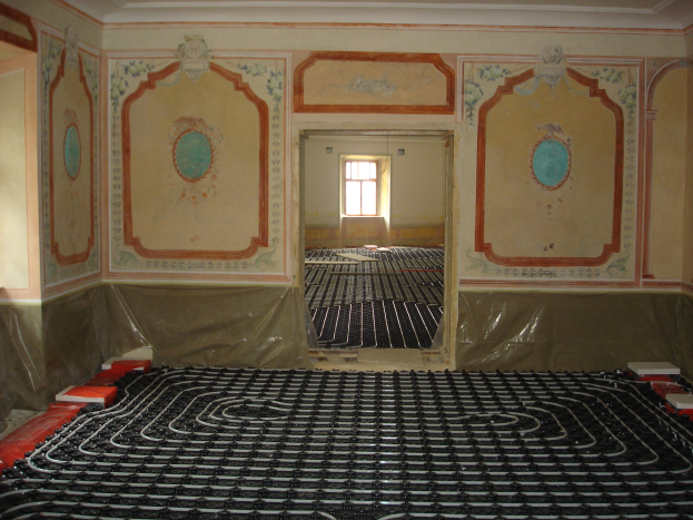 Ein Raum mit einer Fußbodenheizung in der Mitte, umgeben von Wänden und einem Fenster im Hintergrund, mit verschiedenen Gegenständen auf dem Boden, die mit dem Heizungssystem in Zusammenhang stehen.