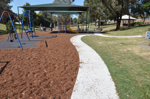 Ein Park mit einem Spielplatz mit Schaukeln und Rutschen, umgeben von Gras, Bäumen, Bänken, Schuppen und einem klaren blauen Himmel.
