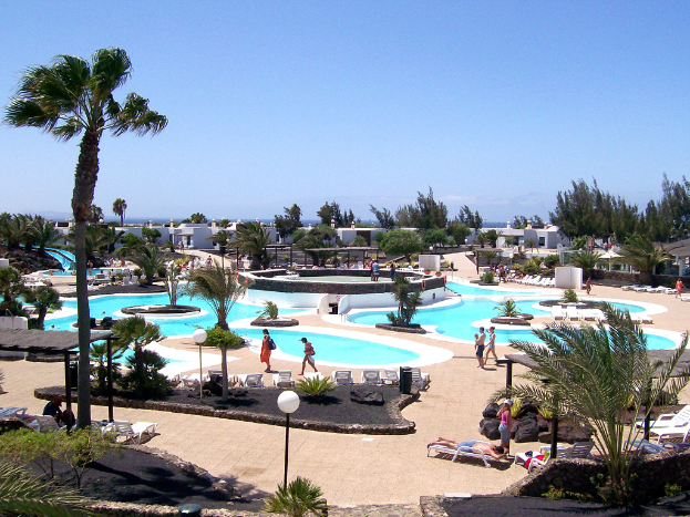 Ein Resort mit Pools umgeben von Bäumen, Menschen zwischen ihnen, Gebäuden im Hintergrund und einem klaren Himmel über ihnen.