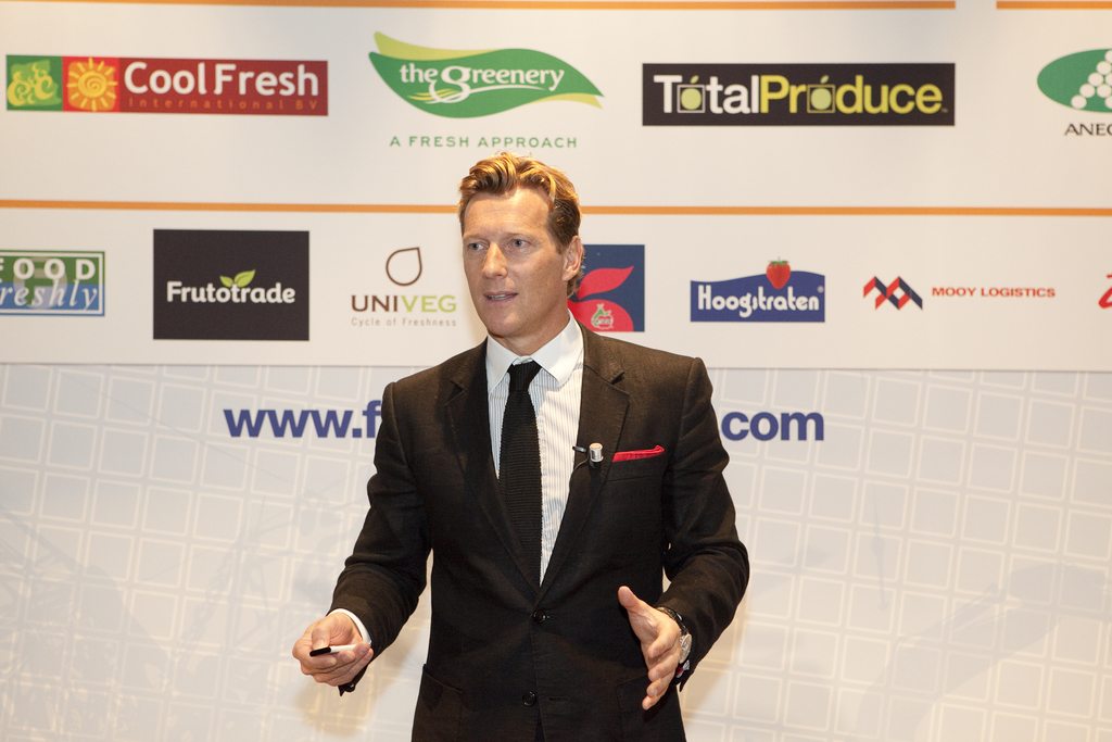 Ein Mann in einem schwarzen Blazer hält ein Objekt und spricht, mit einem weißen Banner und einer weißen Tafel mit organisationalen Logos im Hintergrund.