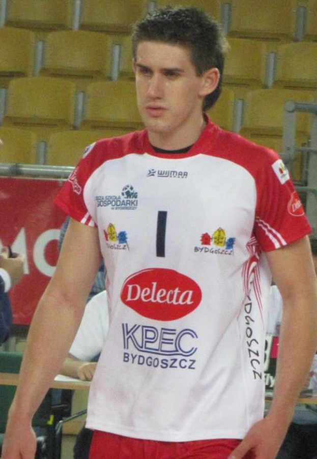 Ein Mann in einem roten und weißen Handballdress steht auf einem Feld, umgeben von Zuschauern, mit einem roten Banner mit der Aufschrift "Krasnoyarsk vs Bydgoszcz" im Hintergrund und sieht entschlossen aus.