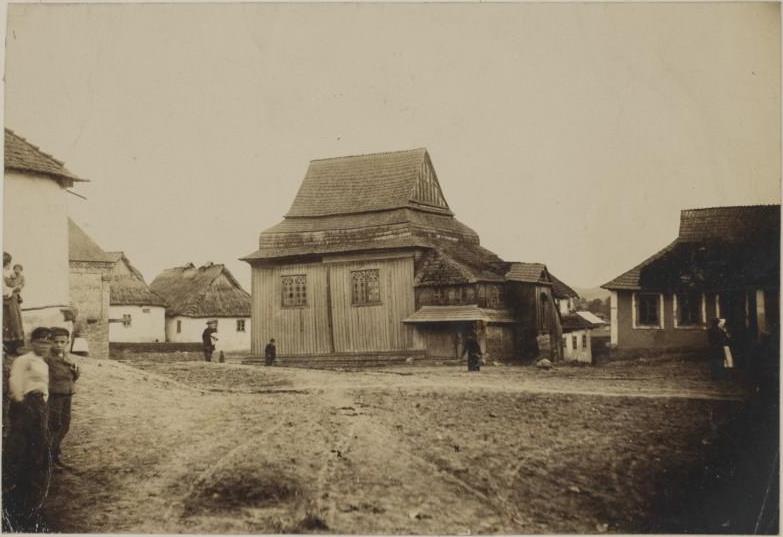 Ein Schwarz-Weiß-Foto einer Gruppe von Menschen, die vor einem hölzernen Haus in Krakau, Polen, stehen, mit anderen Häusern und dem Himmel im Hintergrund.