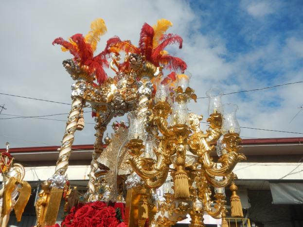 Ein großes goldenes und rotes Festumzugswagen, geschmückt mit Blumen und anderen Dekorationen bei einem Karnevalsumzug, mit einem Gebäude, Strommasten mit Drähten und einem bewölkten Himmel im Hintergrund.