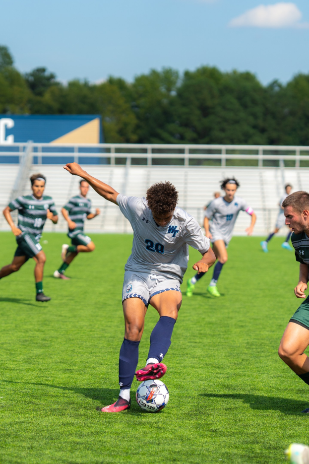 Eine Gruppe junger Männer, die auf einem Feld mit Bäumen und einem klaren blauen Himmel Fußball spielen, wobei ein Spieler Turnschuhe trägt.