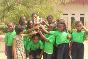 Gruppe von Kindern mit einem Pokal in einer Schulumgebung mit Bäumen und einem Gebäude im Hintergrund.