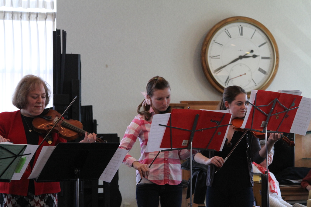 Drei Frauen auf der Bühne mit Geigen stehend und auf Musikständern spielend, während ein sitzendes Publikum zuhört; eine Uhr hängt an der weißen Wand im Hintergrund, und ein weißer Vorhang ist links sichtbar.