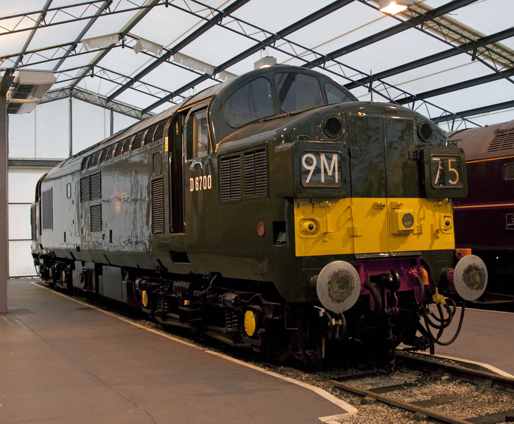 Ein Zug fährt auf den Schienen neben einer Plattform mit einem am Dach befestigten Mast, der Metallstangen mit Lampen trägt.