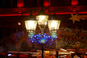 Eine festlich geschmückte Straßenlaterne mit Lichtern und Dekorationen steht nachts in einer Stadt, im Hintergrund ist ein Gebäude zu sehen.