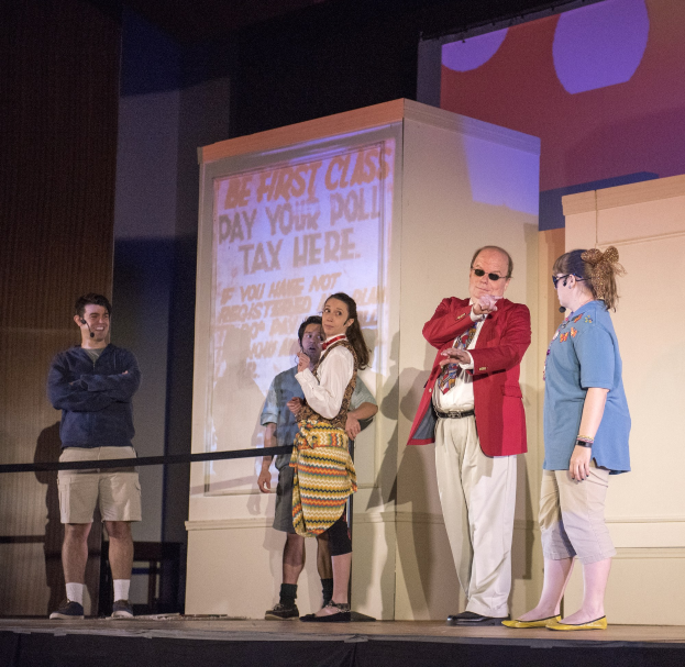 Eine Gruppe von Menschen auf einer Bühne mit einem Mann, der ein Mikrofon hält, ein Display hinter ihnen mit Text, ein Zaun, eine Wand und Deckenleuchten.