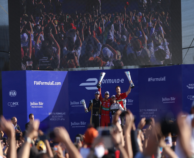 Podium mit einer Gruppe von Menschen, die Tassen halten, FIA Formula E Mexico City Branding im Hintergrund und ein sichtbarer Himmel.