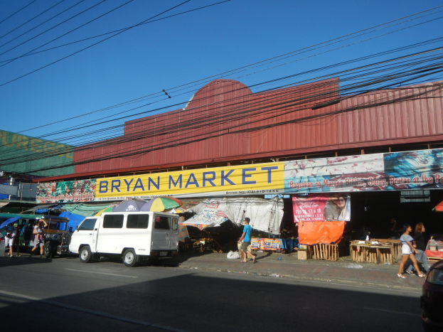 Ein geschäftiger Markt mit Fahrzeugen, Fußgängern, Marktständen, Bannern, Schirmen, hölzernen Gegenständen und einem "Bryan Market"-Schild im Hintergrund unter einem Himmel mit Stromleitungen.