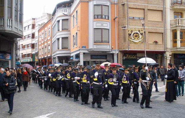 Eine Gruppe von Menschen, die mit Schirmen auf der Straße gehen, einige halten Musikinstrumente und andere tragen Taschen, mit Gebäuden, Laternenmasten und Namensschildern im Hintergrund und einem sichtbaren Himmel, während eines Ehrenzuges.