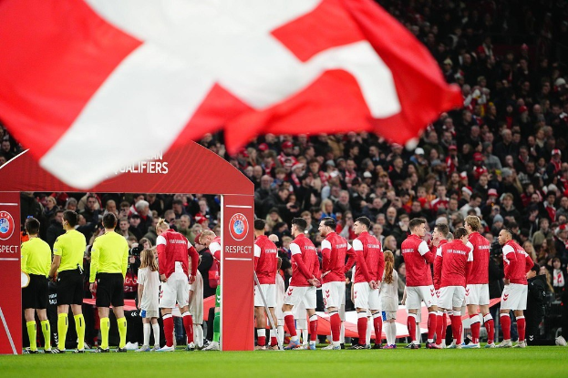 Gruppe von Menschen auf einem Fuballfeld mit einer roten und weißen Flagge im Vordergrund, einem Bogen mit der Aufschrift "Bayern Munich vs Bayern Munich Betting Tips & Preview" im Hintergrund und einer großen Menge im Stadion.