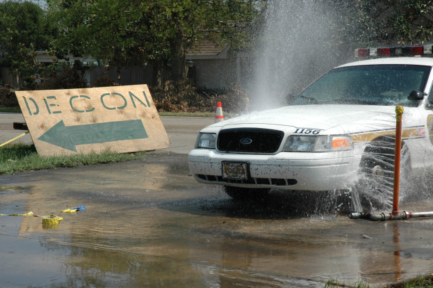 Ein Polizeiwagen fährt durch eine Pfütze, mit einer Tafel, einem Verkehrskegel und anderen Gegenständen am Boden, vor einer Kulisse aus Bäumen, Gebäuden und einem klaren blauen Himmel.