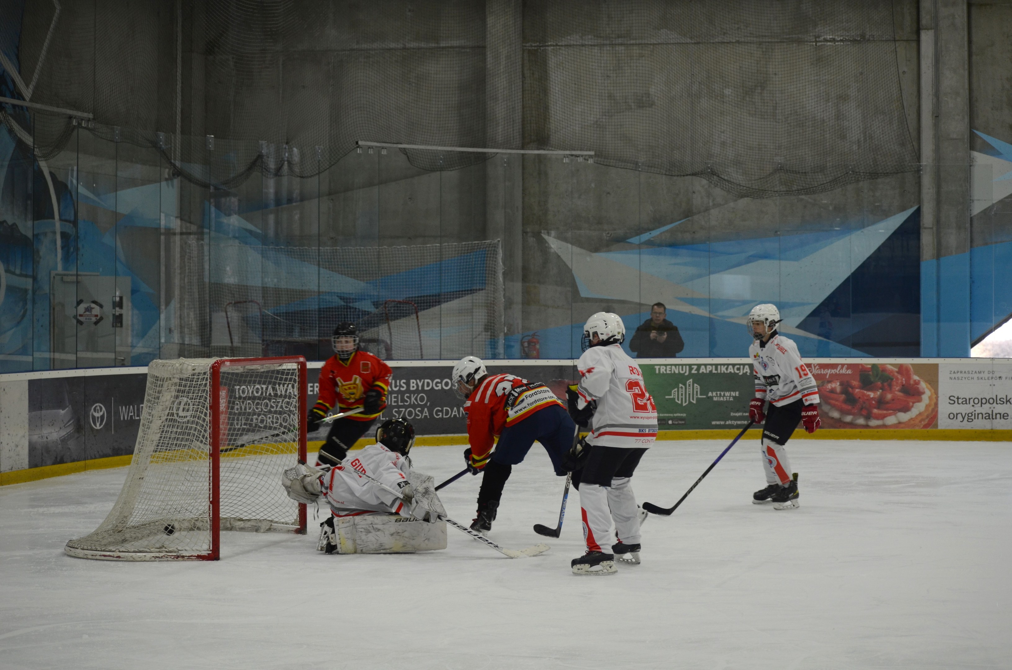 Eishockey-Spieler in Helmen und Uniformen konkurrieren in der Nähe eines Torpfostens auf einer Indoor-Eisbahn, mit Bannern und einer Wand im Hintergrund.