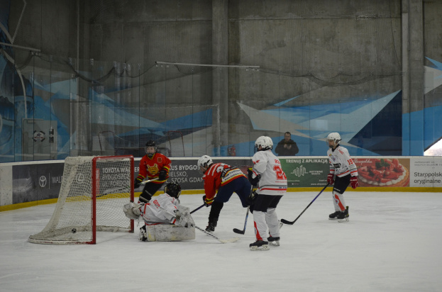 Eishockey-Spieler in Helmen und Uniformen konkurrieren in der Nähe eines Torpfostens auf einer Indoor-Eisbahn, mit Bannern und einer Wand im Hintergrund.
