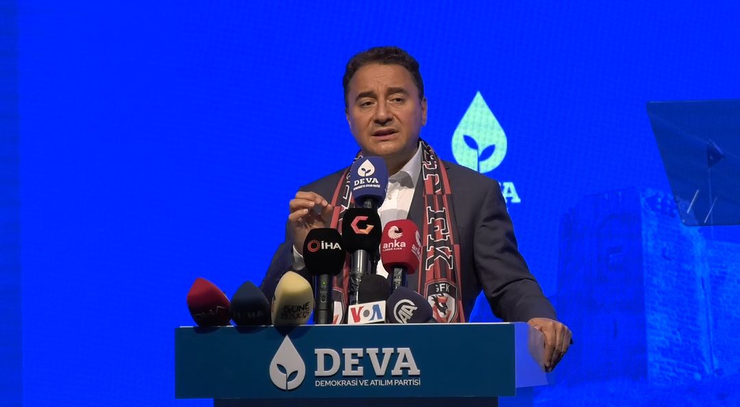 Ein Mann in Mantel und Schal steht an einem Podium mit Mikrofonen, ein Schild mit der Aufschrift "deva ceo sagt, es ist keine gute Idee für die Zukunft", ein Objekt und ein Bildschirm im Hintergrund.