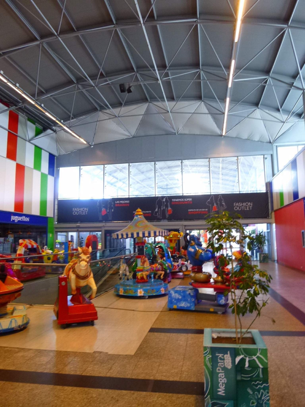 Großer Indoor-Vergnügungspark in einem Einkaufszentrum mit verschiedenen Fahrgeschäften, einem Topf mit einer Pflanze, verstreuten Spielzeugen, Informationsschildern, Deckenleuchten und Metallträgern.
