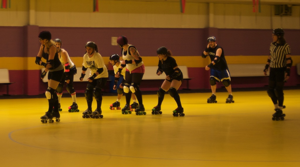 Menschen in Kleidung skaten auf einem Boden mit Bänken vor einer Wand.
