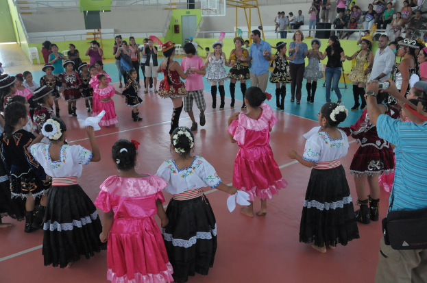 Eine Gruppe von Kindern in Kostümen tanzt in der Mitte des Bildes, während das Publikum von oben rechts zusieht.