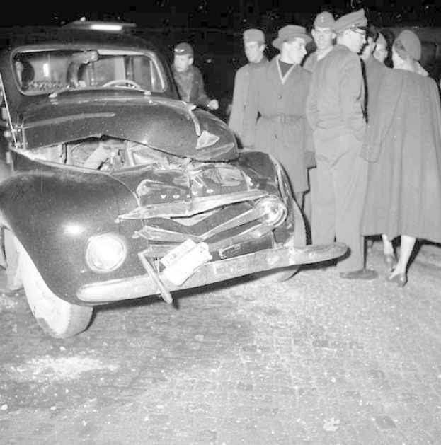 Ein Schwarz-Weiß-Bild eines schwer beschädigten Autos am Straßenrand mit einer Gruppe von Menschen, die es inspizieren.