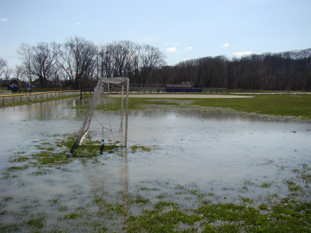 Ein Fussballtor steht inmitten eines überfluteten Feldes, umgeben von Gras, einem Zaun, Bäumen, Häusern und einem bewölkten Himmel.