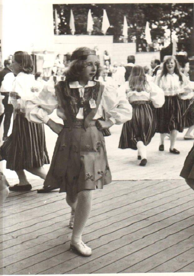 Eine Gruppe von Frauen in traditioneller bayrischer Tracht tanzt auf einem Holzboden, mit einer Wand, Bäumen und anderen Gegenständen im Hintergrund. Text ist am unteren Rand des Bildes sichtbar.
