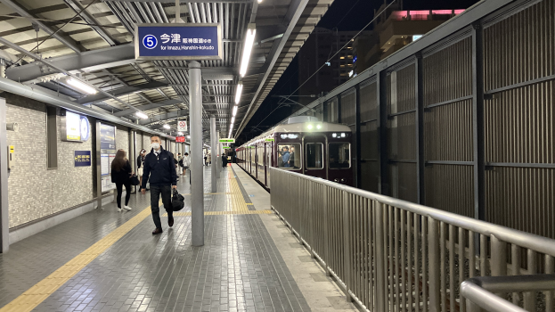 Ein Zug fährt auf einem Bahnsteig ein, mit Menschen in der Nähe, einem Geländer auf der rechten Seite, Gebäuden im Hintergrund, Bahnsteiglütung und einer Abfahrtsanzeige.