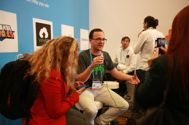 Ein Interview findet statt mit Menschen, die Mikrofone und eine Kamera halten, auf der linken Seite ist ein Banner zu sehen und es gibt Personen im Vordergrund und in der Mitte, mit einem klaren Hintergrund.