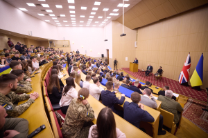 Eine große Gruppe von Menschen an Tischen in einem Raum mit Flaggen auf der rechten Seite, eine Wand im Hintergrund, Papiere auf den Tischen, einige Stehende vor einem Podium mit Mikrofon, Deckenleuchten und ein Projektor.