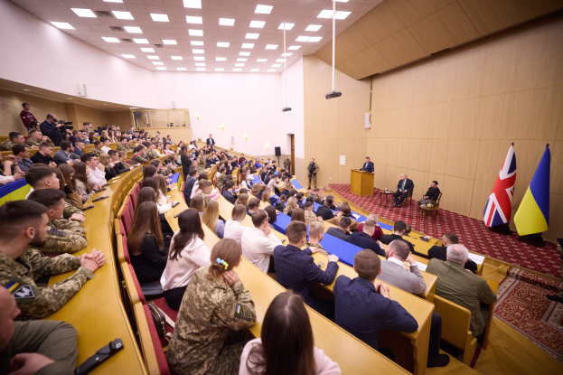 Eine große Gruppe von Menschen an Tischen in einem Raum mit Flaggen auf der rechten Seite, eine Wand im Hintergrund, Papiere auf den Tischen, einige Stehende vor einem Podium mit Mikrofon, Deckenleuchten und ein Projektor.