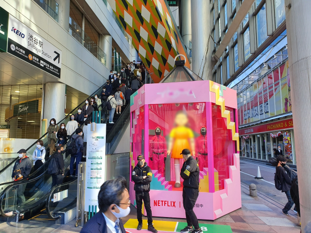 Eine Gruppe von Menschen steht vor einem Gebäude mit Glaswänden, Säulen und Texttafeln, einige tragen Masken, während eine Rolltreppe mit Menschen in der Nähe nach oben führt; eine pinkfarbene Box in der Mitte zeigt Schaufensterpuppen und trägt die Aufschrift "Netflix' neuester Netflix-Stand in Tokyo."