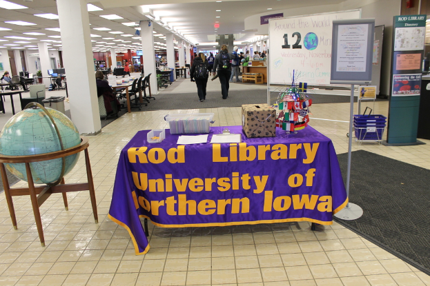 Eine Ausstellung mit einem Tisch, auf dem verschiedene Landesflaggen, eine Box, Papiere und ein Banner liegen, eine Kugel links daneben, eine Anschlagtafel rechts daneben mit ein paar Leuten daneben und mehreren Tischen und Stühlen links.