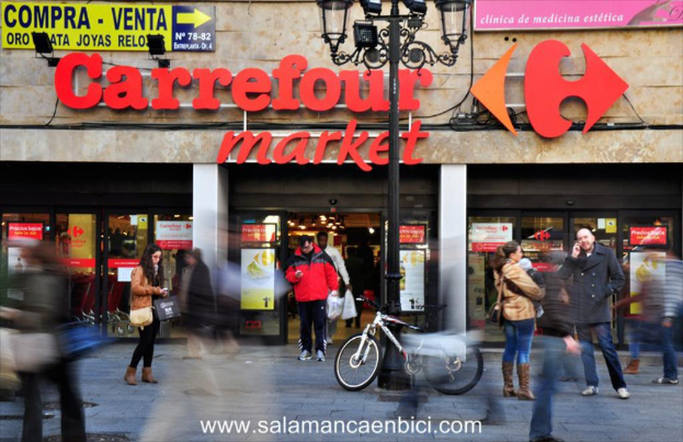 Ein belebter Einkaufsmarkt mit Menschen vor einem Einkaufszentrum, einem schwarzen Pfahl mit Lichtern, einem Fahrrad daneben und einer Richtungstafel an der Wand.