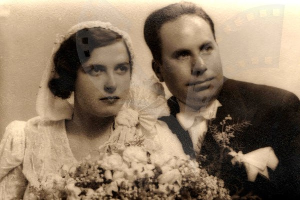 Ein Schwarz-Weiß-Foto eines strahlenden Mannes und einer Frau an ihrem Hochzeitstag, die Frau in einem weißen Kleid mit einem Blumenstrauß, der Mann neben ihr in feierlicher Kleidung.