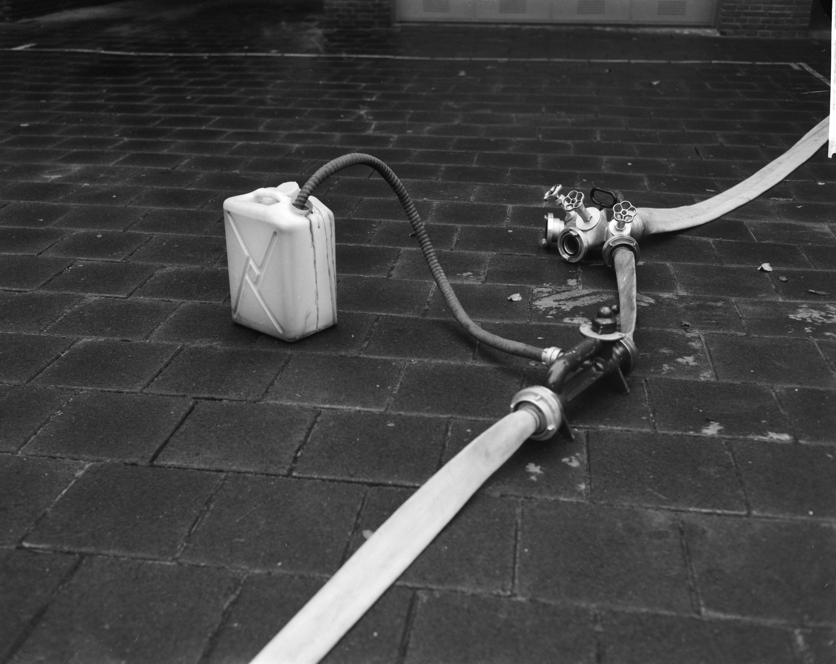 Schwarz-weiß-Foto eines Gasbehälters mit einem Schlauch, der vor einem Gebäude mit einer Tür im Hintergrund auf dem Boden steht.